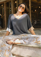 A woman wearing a dark gray V-neck top with beige rolled cuffs and wide-leg beige paisley pants, sitting on a wooden ledge in front of a slatted wooden wall.