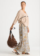 A woman modeling a beige draped top and a beige-and-white paisley print maxi skirt, holding a brown woven tote bag against a light gray studio background.