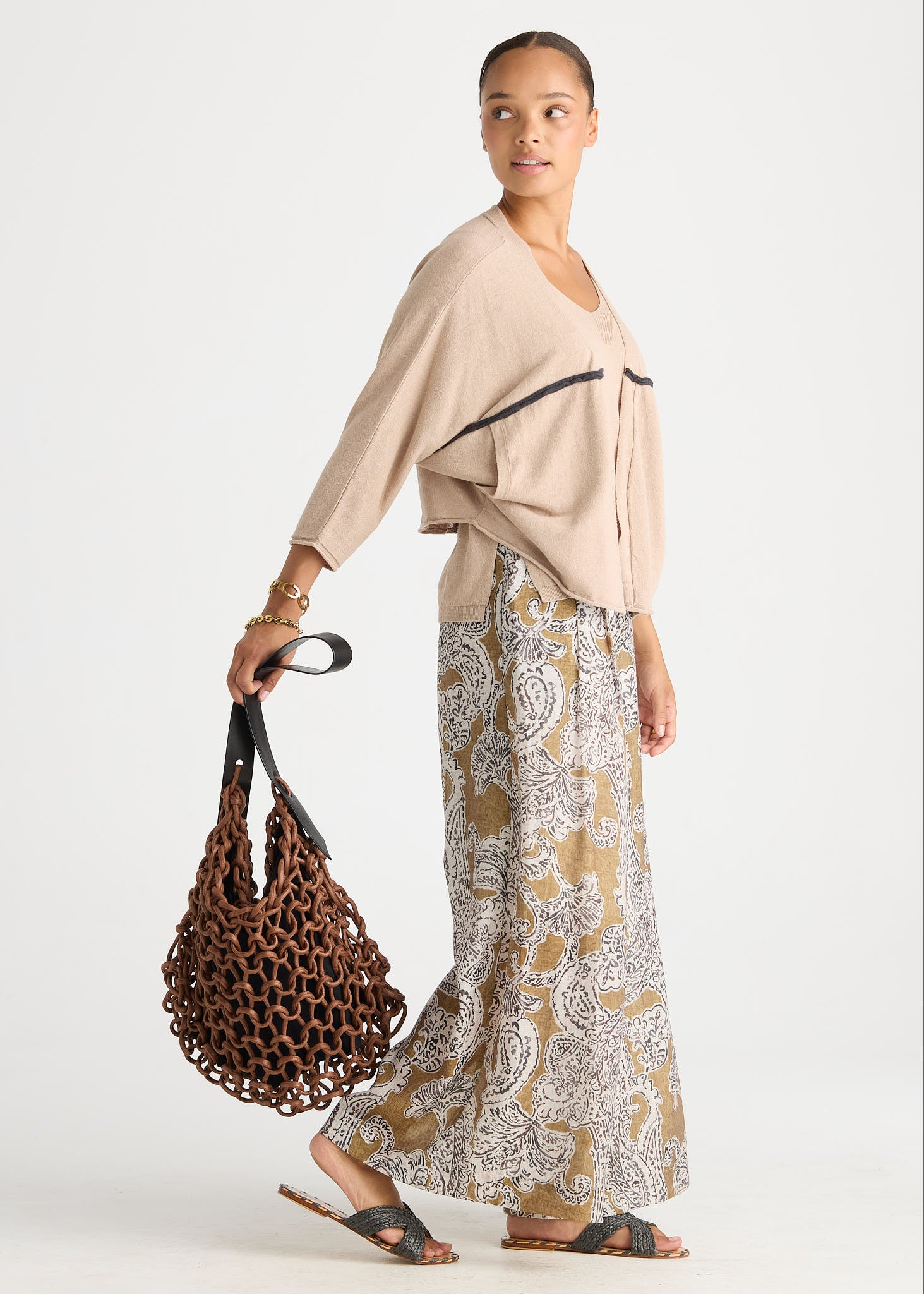 A woman modeling a beige draped top and a beige-and-white paisley print maxi skirt, holding a brown woven tote bag against a light gray studio background.