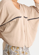 A close-up of a person wearing a light beige cropped cardigan with dark contrast stitching and a V-neck, paired with patterned pants.