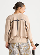 A woman shown from the back wearing a beige knit top with black contrast stitching forming a cross design on the back, paired with patterned pants against a light gray studio background.