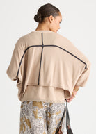 A woman shown from the back wearing a beige knit poncho-style top with black contrast piping and a geometric cross design on the back, paired with patterned pants against a light gray studio background.