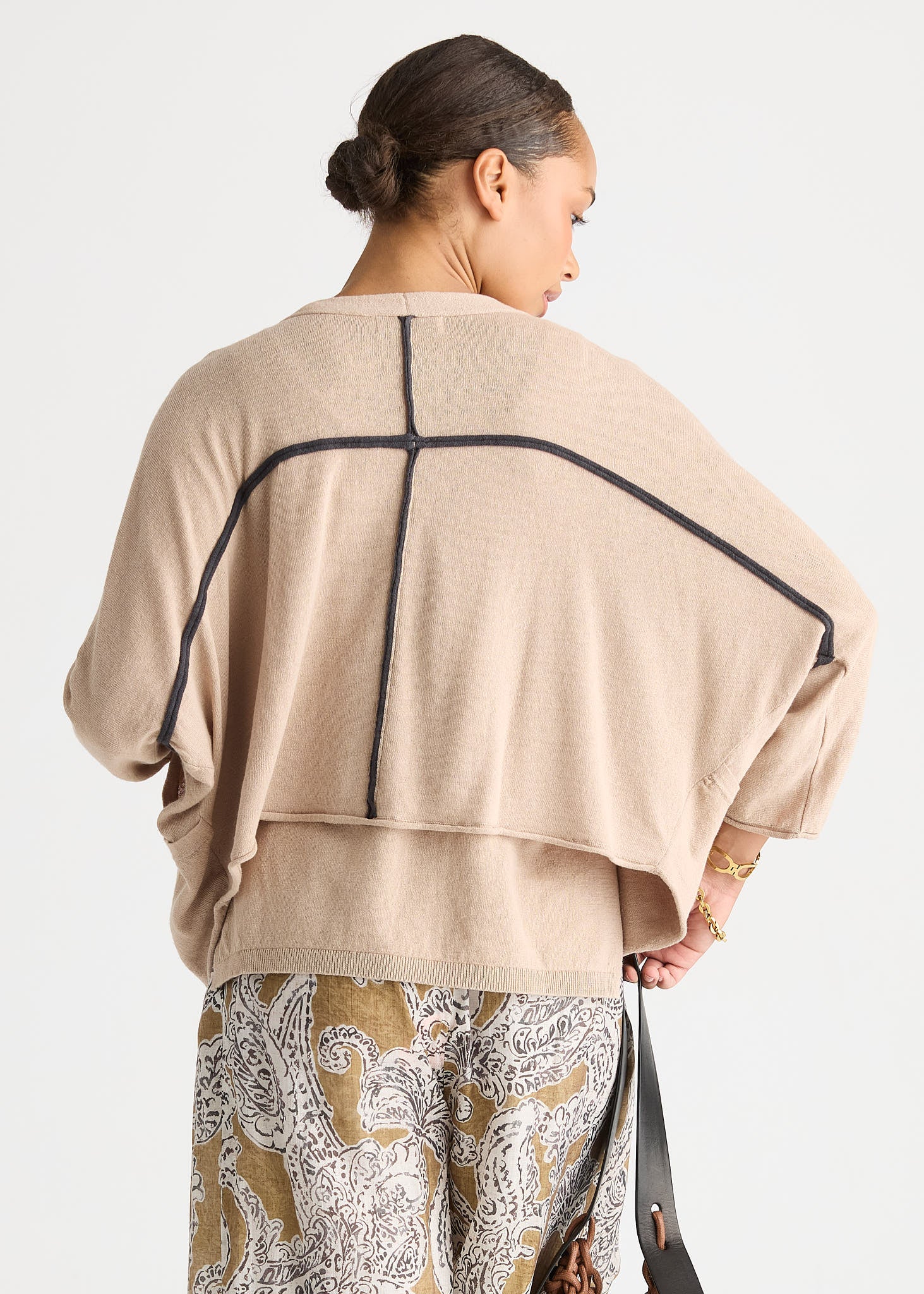 A woman shown from the back wearing a beige knit poncho-style top with black contrast piping and a geometric cross design on the back, paired with patterned pants against a light gray studio background.