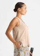 A woman shown in side profile wearing a light beige sleeveless V-neck top and patterned pants, standing against a plain light gray studio background.