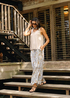 A woman wearing a sleeveless cream top and patterned beige-and-white wide-leg pants walks down wooden stairs in front of a rustic wooden building.