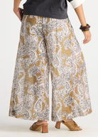 A person wearing wide-leg cropped pants with a beige, white, and gray paisley pattern, styled with a dark gray top and tan heeled mule sandals against a plain white studio background.