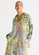 A model wearing a long-sleeved, button-up shirt dress with a vibrant multicolor patchwork print of green, yellow, red, and blue botanical patterns, styled with a chunky green beaded necklace against a plain white background.