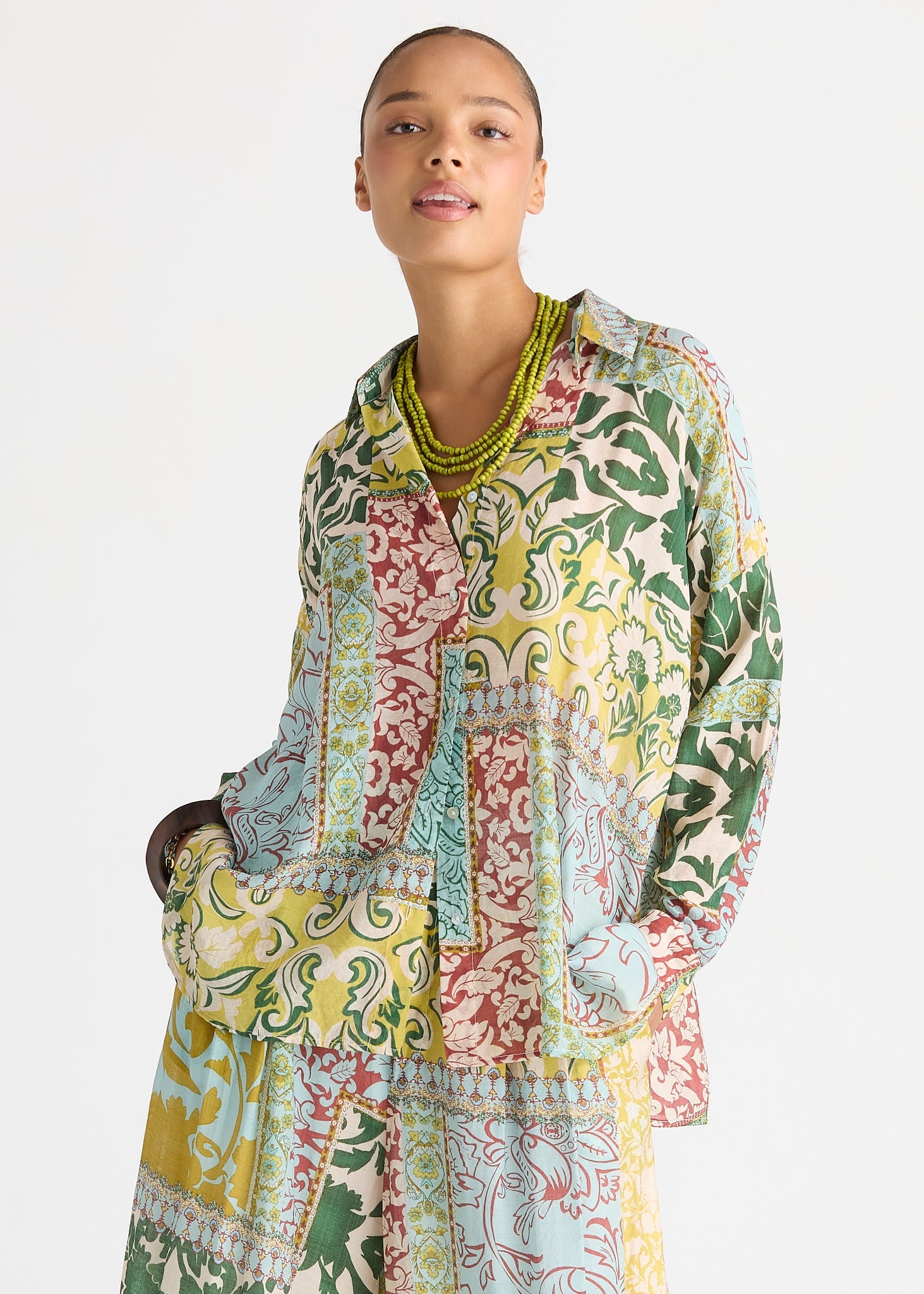 A model wearing a long-sleeved, button-up shirt dress with a vibrant multicolor patchwork print of green, yellow, red, and blue botanical patterns, styled with a chunky green beaded necklace against a plain white background.