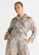 A woman wearing a long-sleeve collared wrap-style dress with a brown, white, and black paisley pattern, posed against a light gray studio background.