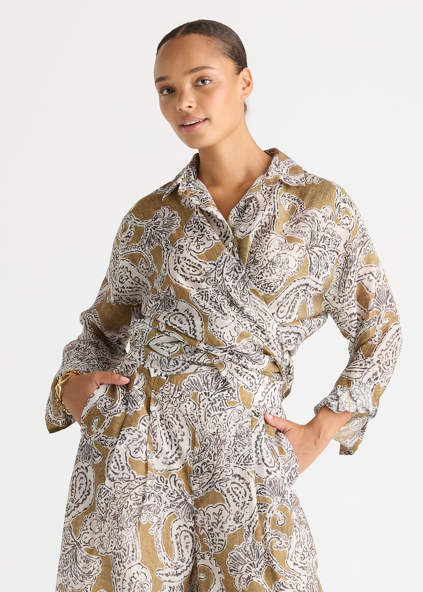 A woman wearing a long-sleeve collared wrap-style dress with a brown, white, and black paisley pattern, posed against a light gray studio background.