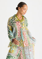 A woman wearing a long-sleeve patchwork-print tunic top with green, yellow, pink, and blue patterns, styled with a chunky green beaded necklace against a light gray background.