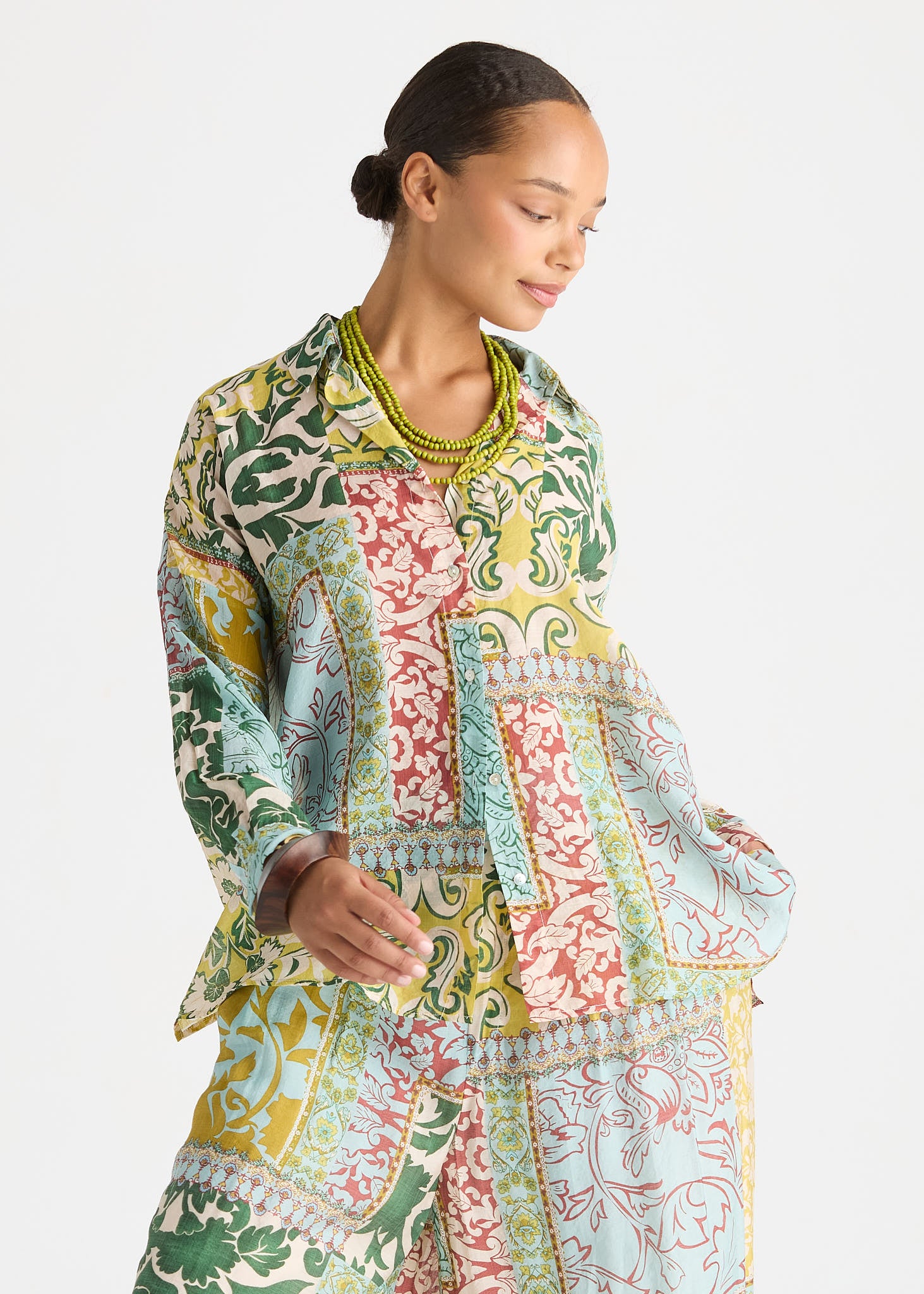 A woman wearing a long-sleeve patchwork-print tunic top with green, yellow, pink, and blue patterns, styled with a chunky green beaded necklace against a light gray background.