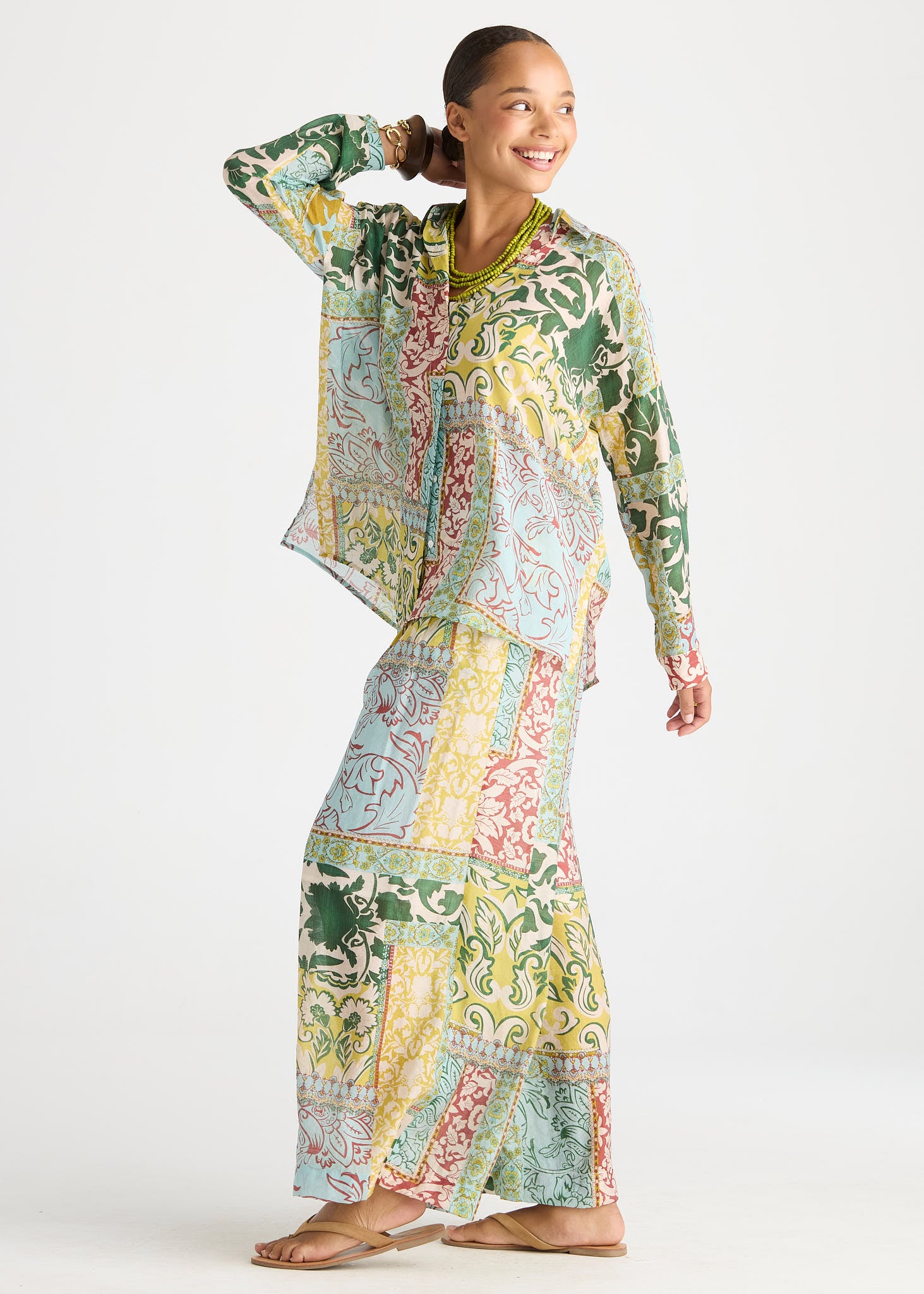 A woman modeling a matching two-piece printed lounge set consisting of a long-sleeve button-up shirt and a long maxi skirt, featuring a multicolor patchwork paisley pattern in green, yellow, pink, and blue on a white background.