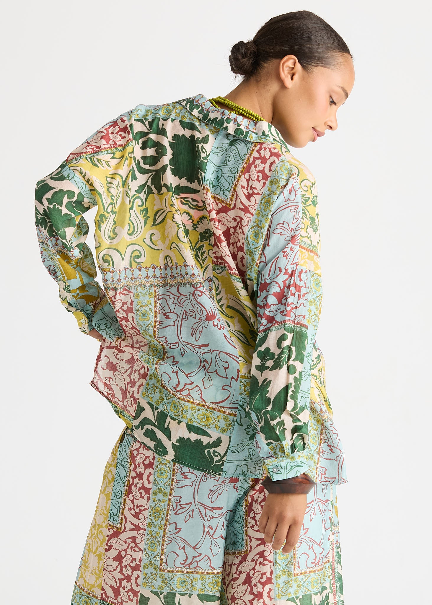 A woman shown from the back wearing a loose, long-sleeve patchwork-print top and matching bottoms. The fabric features a multicolor patchwork design with green, yellow, red, and light blue patterns on a cream base, set against a plain light gray studio background.