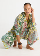 A woman crouches barefoot on a white studio floor wearing a loose, patterned kaftan-style dress with green, yellow, red, and blue botanical motifs.