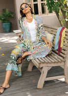 A woman wearing a matching tropical print lounge set consisting of a lightweight open-front jacket and wide-leg pants, sitting on a wooden bench on a deck.
