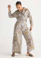 A woman modeling a long-sleeve, collared jumpsuit with a wide-leg pant and a brown, beige, and white paisley pattern, paired with dark brown slide sandals against a plain white studio background.