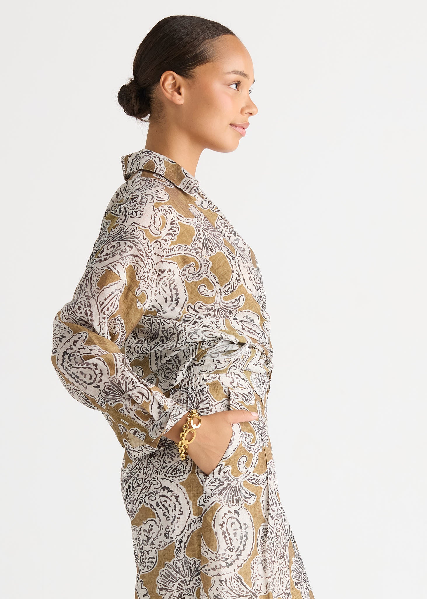 A woman in side profile wearing a long-sleeve collared shirt dress with a beige, white, and gray paisley pattern, set against a plain light gray background.