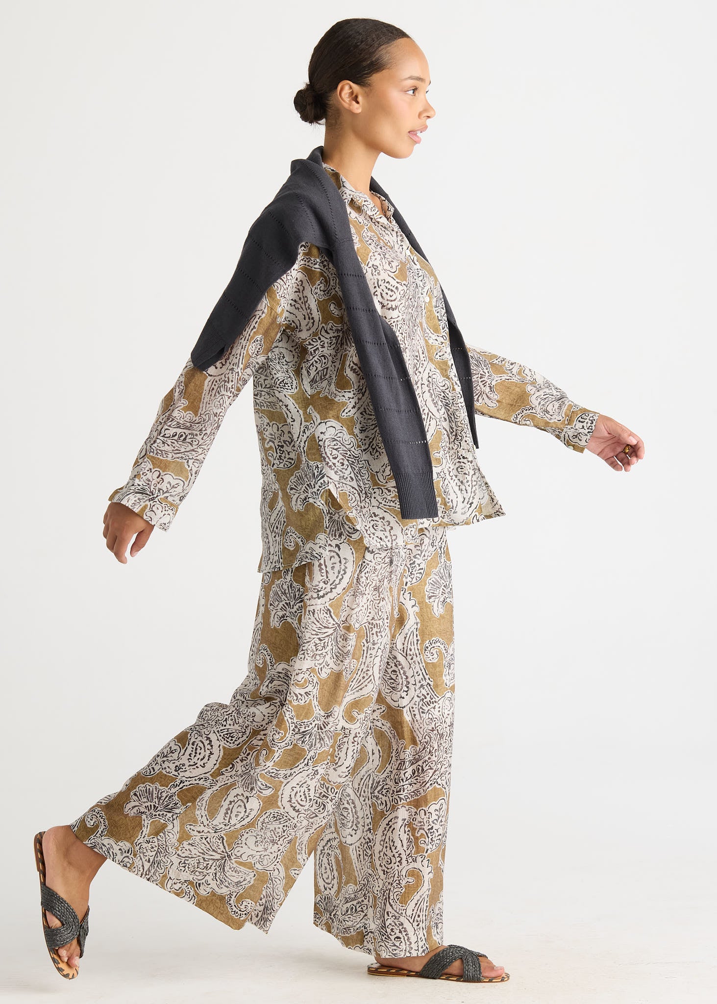 A woman modeling a matching two-piece outfit with a beige, white, and gray paisley pattern, featuring a long-sleeve top and wide-leg pants, styled with a black draped jacket and dark sandals against a plain white background.