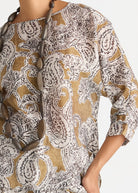 A close-up of a woman wearing a loose, three-quarter sleeve top with an intricate paisley pattern in brown, beige, and white. A long brown beaded necklace is draped over the chest.