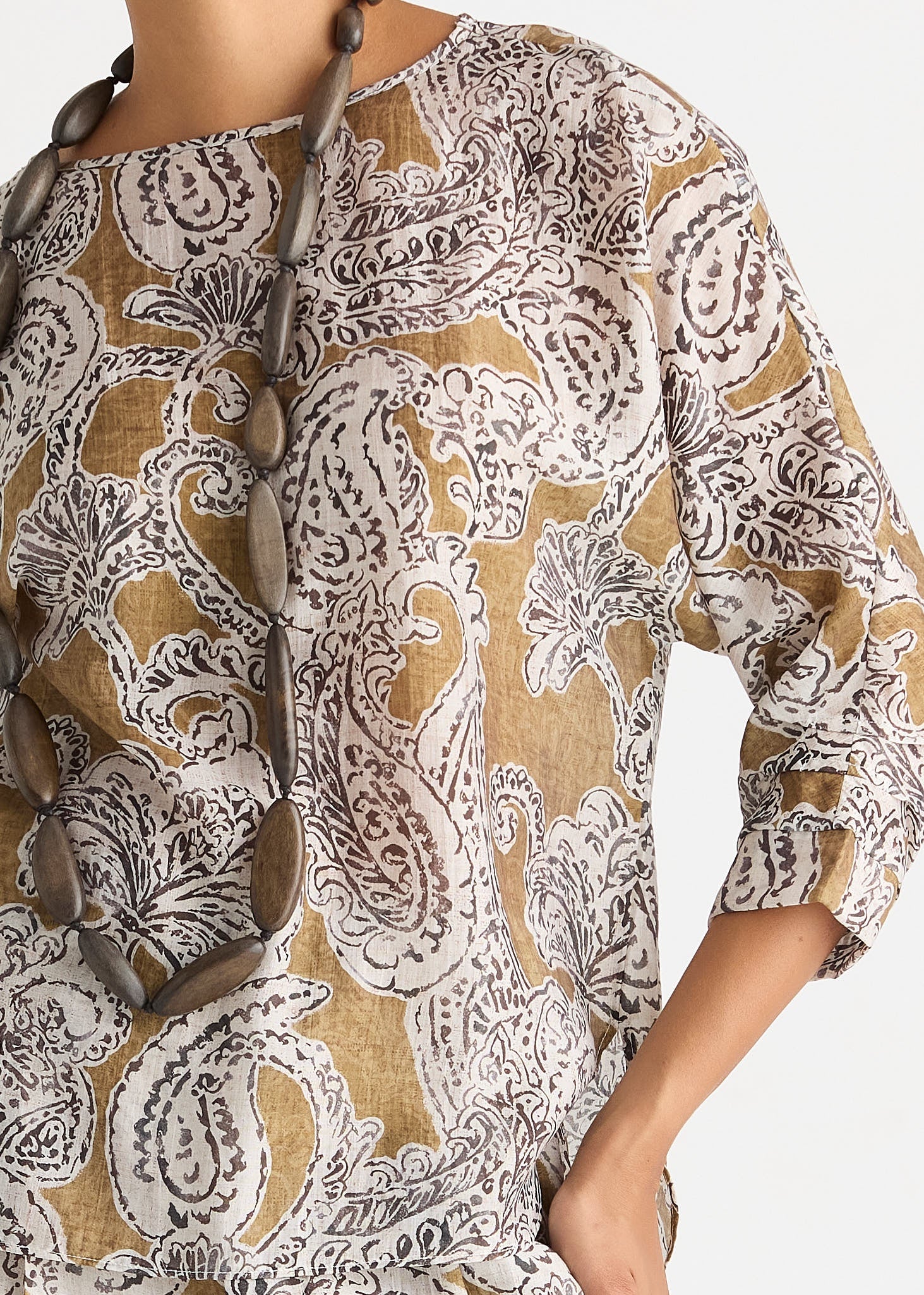 A close-up of a woman wearing a loose, three-quarter sleeve top with an intricate paisley pattern in brown, beige, and white. A long brown beaded necklace is draped over the chest.