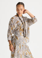 A woman modeling a matching two-piece set featuring a beige and white paisley pattern, including a long-sleeve top and a pleated skirt, styled with a long dark belt and gold jewelry against a plain light gray background.