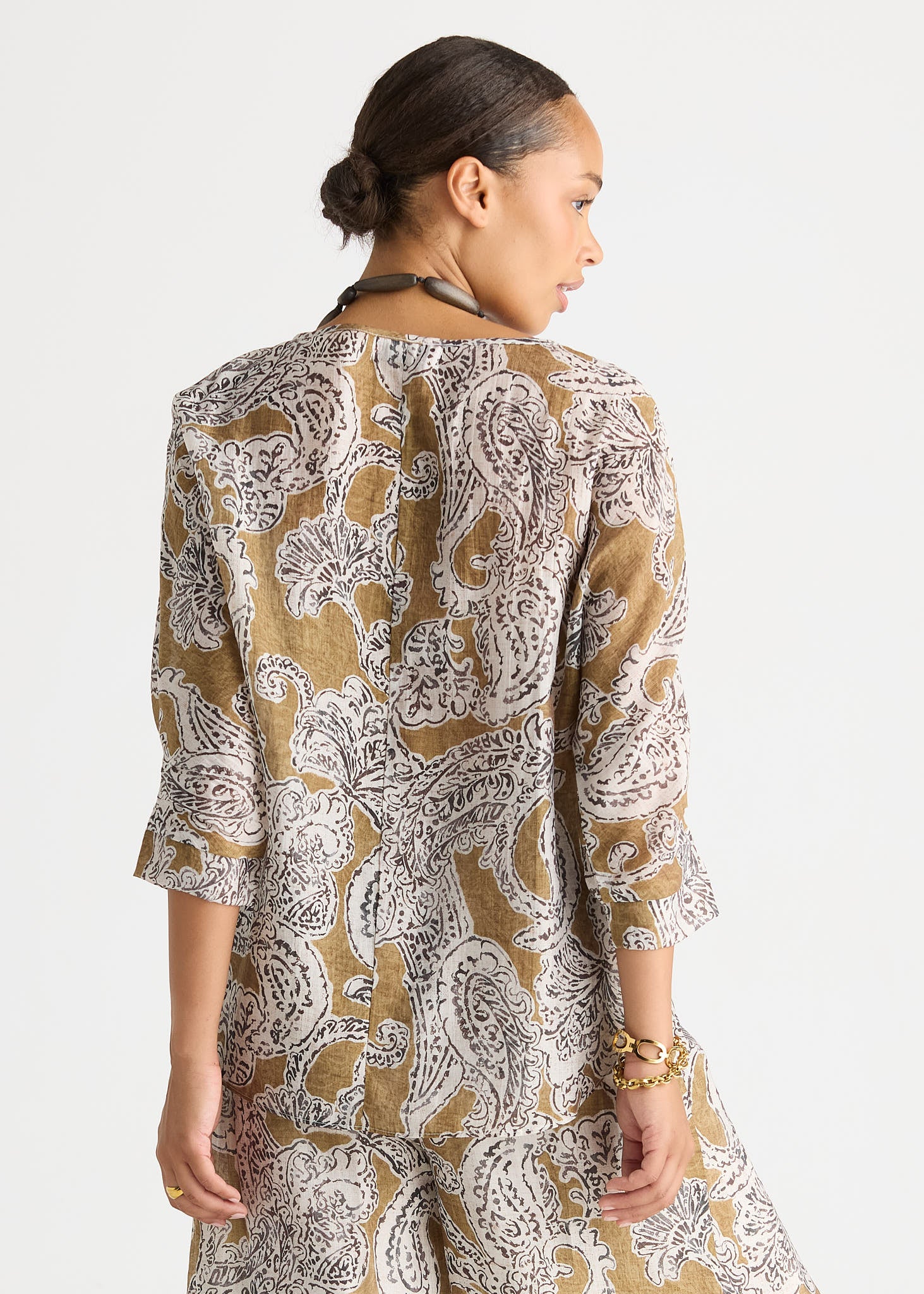 A woman shown from the back wearing a loose, three-quarter sleeve tunic or dress with an allover paisley print in tan, white, and gray on a plain light gray studio background.