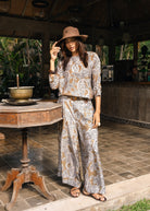 A woman stands in a rustic outdoor patio setting wearing a matching beige and brown paisley print two-piece set with a wide-brimmed hat.