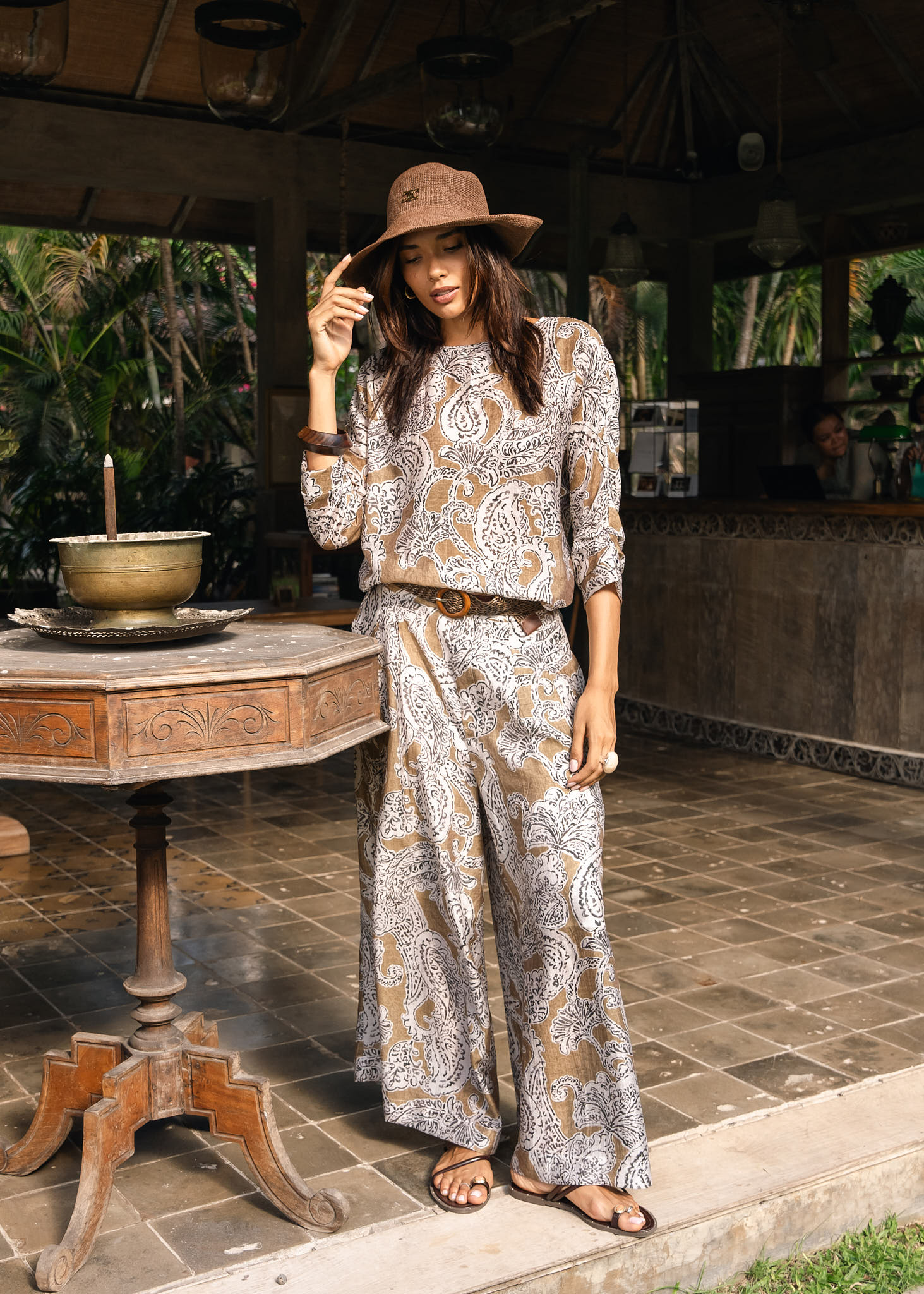 A woman stands in a rustic outdoor patio setting wearing a matching beige and brown paisley print two-piece set with a wide-brimmed hat.