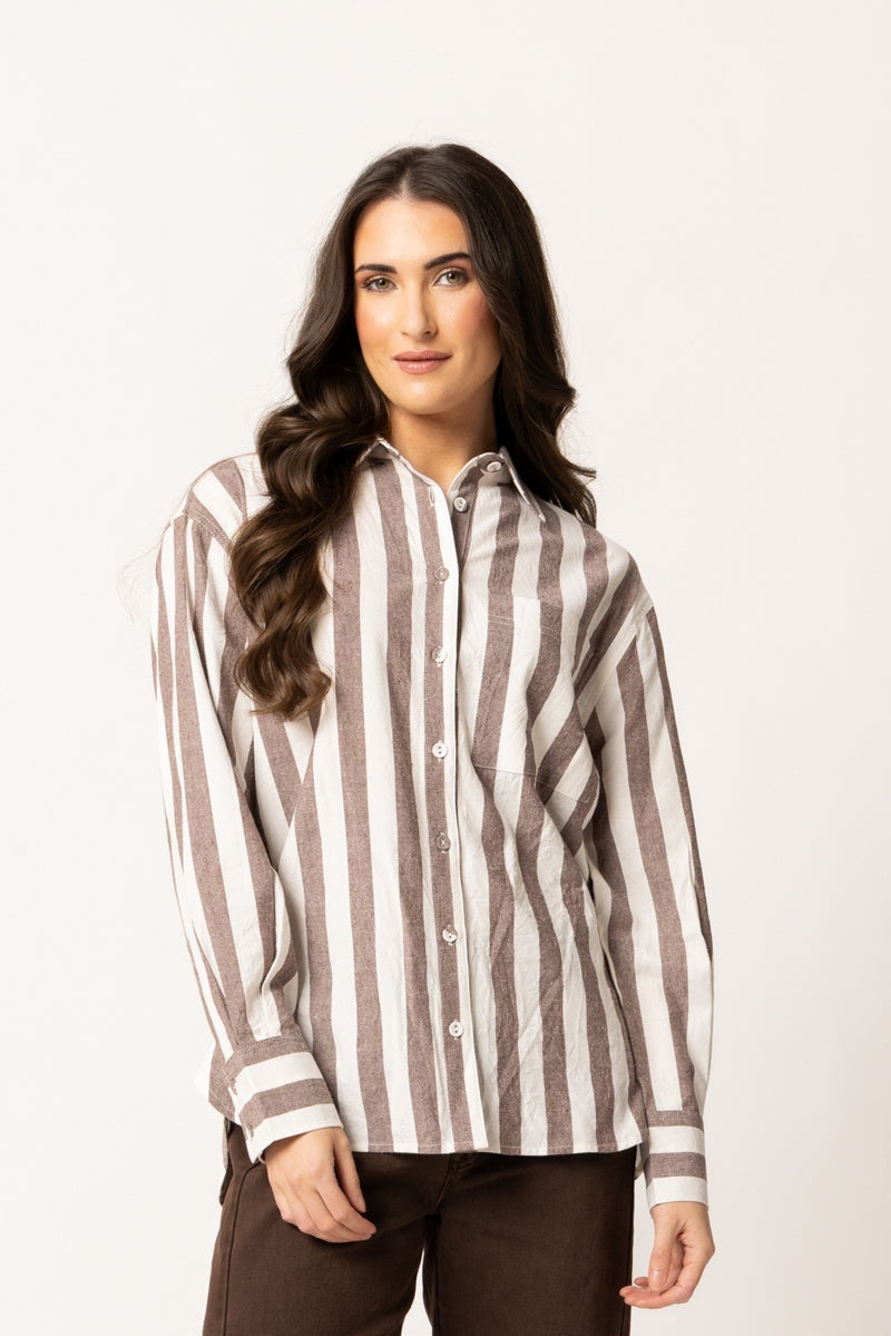 A woman modeling a long-sleeve button-up shirt with vertical brown and off-white stripes, paired with dark brown pants, against a plain light background.