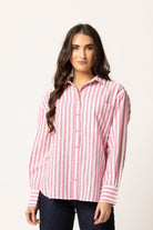 A woman modeling a pink and white vertically striped button-down shirt with dark blue jeans, photographed against a plain off-white studio background.