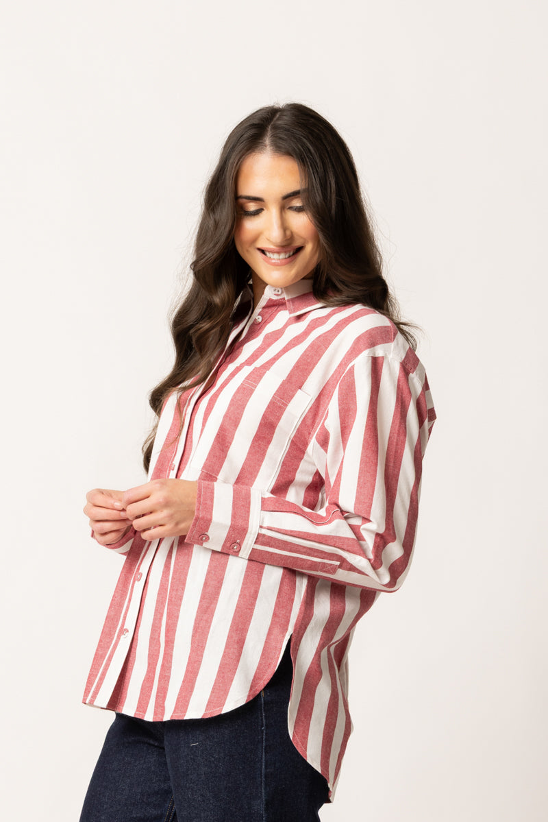 A woman modeling a long-sleeve button-up shirt with vertical pink and white stripes, paired with dark blue jeans, against a plain off-white studio background.