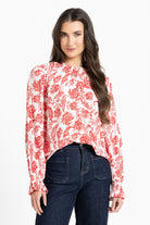 A woman modeling a long-sleeve blouse with a red floral and paisley print on a white background, paired with dark blue jeans, against a plain white studio background.