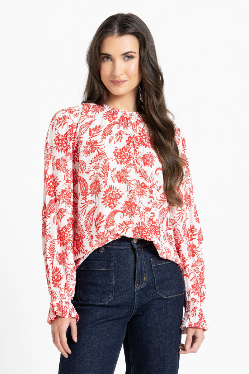 A woman modeling a long-sleeve blouse with a red floral and paisley print on a white background, paired with dark blue jeans, against a plain white studio background.
