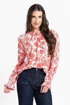 A woman modeling a long-sleeve blouse with a red and white floral pattern, paired with dark blue jeans, against a plain white background.