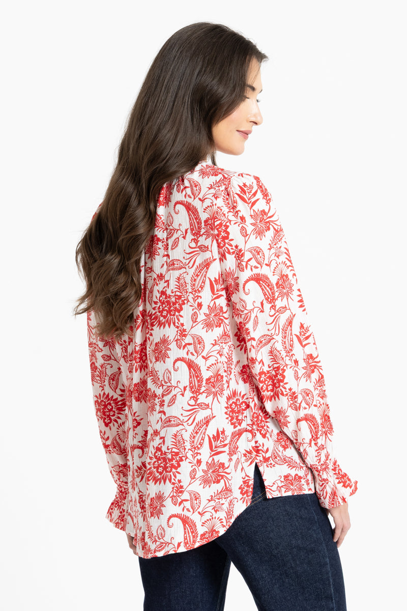 A woman shown from the back wearing a white long-sleeve blouse with a red floral and paisley print, paired with dark blue jeans, against a plain white background.