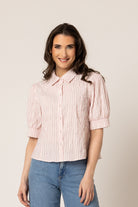 A woman wearing a light pink and white vertically striped short-sleeve blouse with puff sleeves, paired with blue jeans, standing against a plain beige studio background.