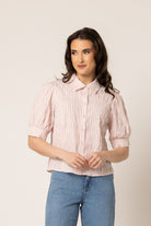 A woman wearing a light pink and white vertically striped short-sleeve blouse with puff sleeves, paired with blue jeans, standing against a plain beige studio background.