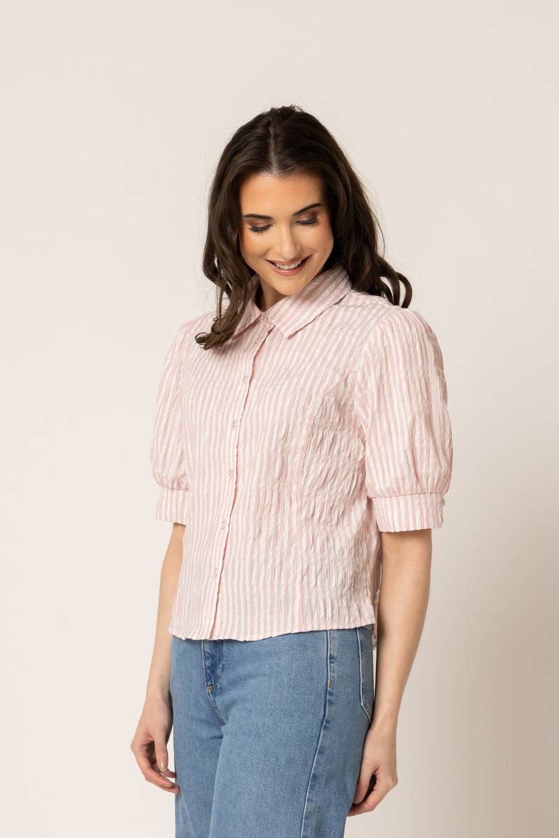 A woman wearing a light pink and white vertically striped short-sleeve blouse with puff sleeves, paired with blue jeans, standing against a plain beige studio background.