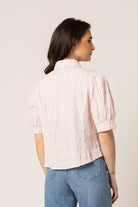A woman wearing a light pink and white vertically striped short-sleeve blouse with puff sleeves, paired with blue jeans, standing against a plain beige studio background.