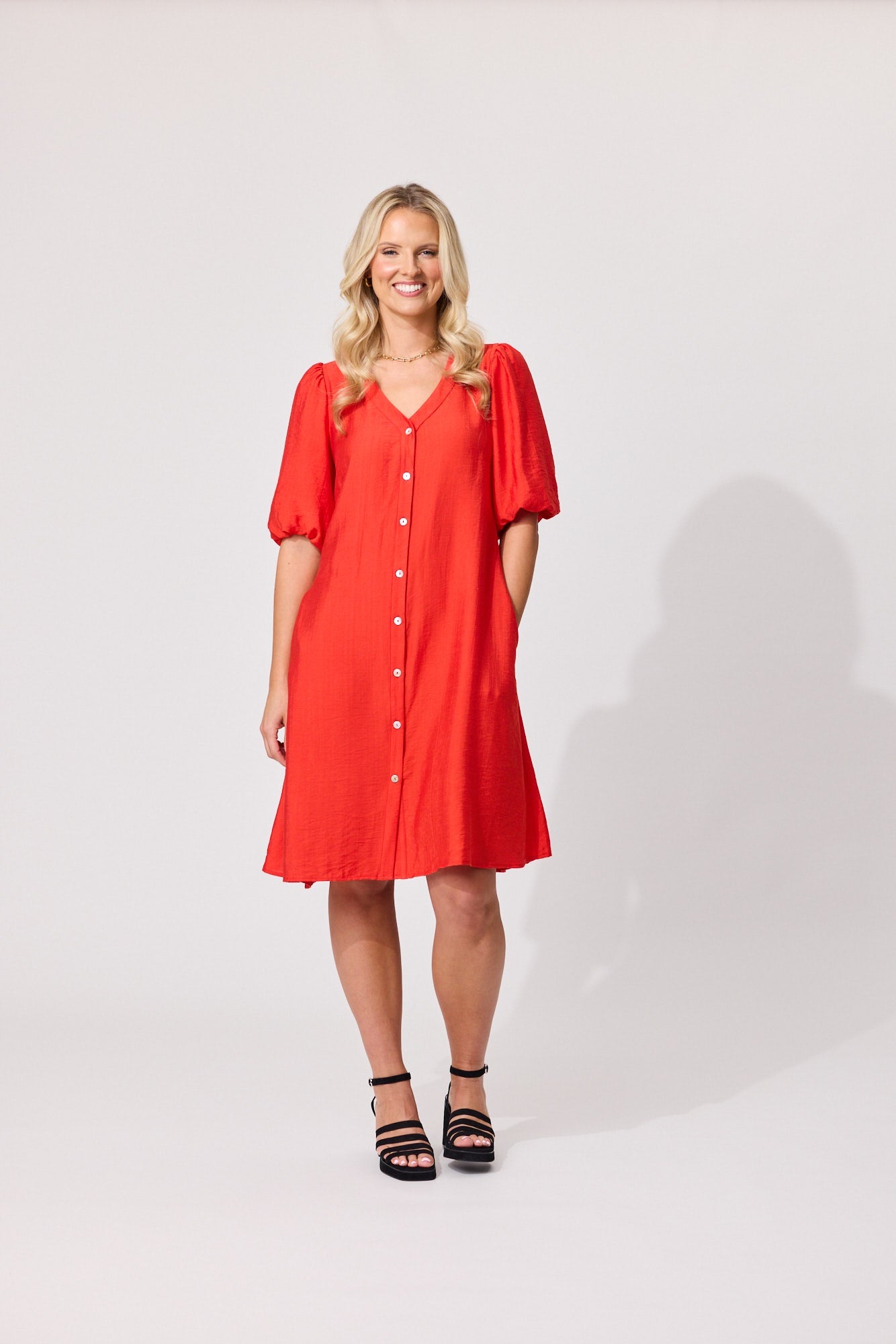 A woman modeling a bright red short-sleeve button-front dress with puff sleeves, paired with black strappy heels, against a light gray studio background.