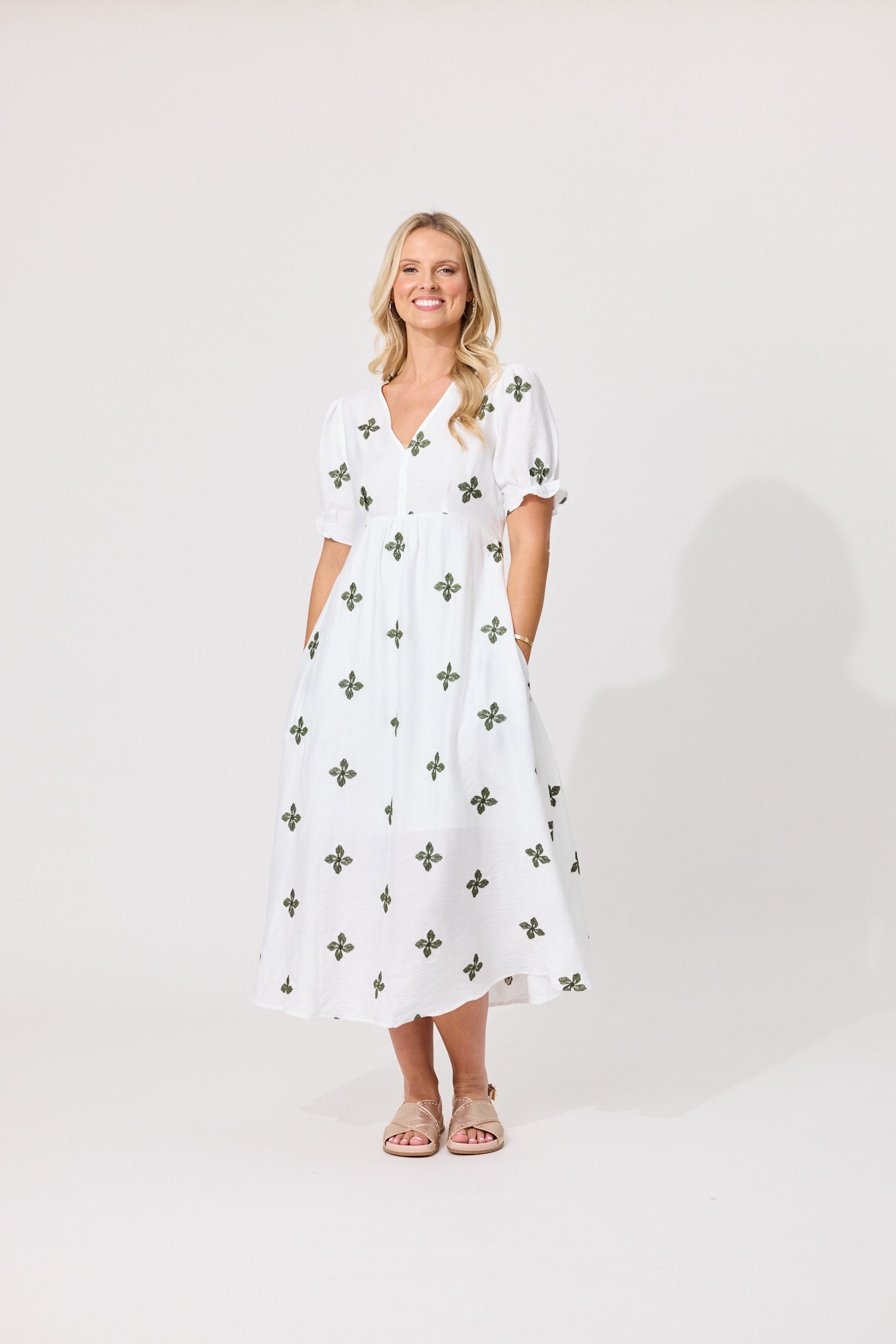 A woman modeling a white midi dress with a green cross pattern, standing against a plain light gray studio background.