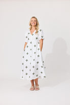 A woman modeling a white midi dress with a green cross pattern, standing against a plain light gray studio background.