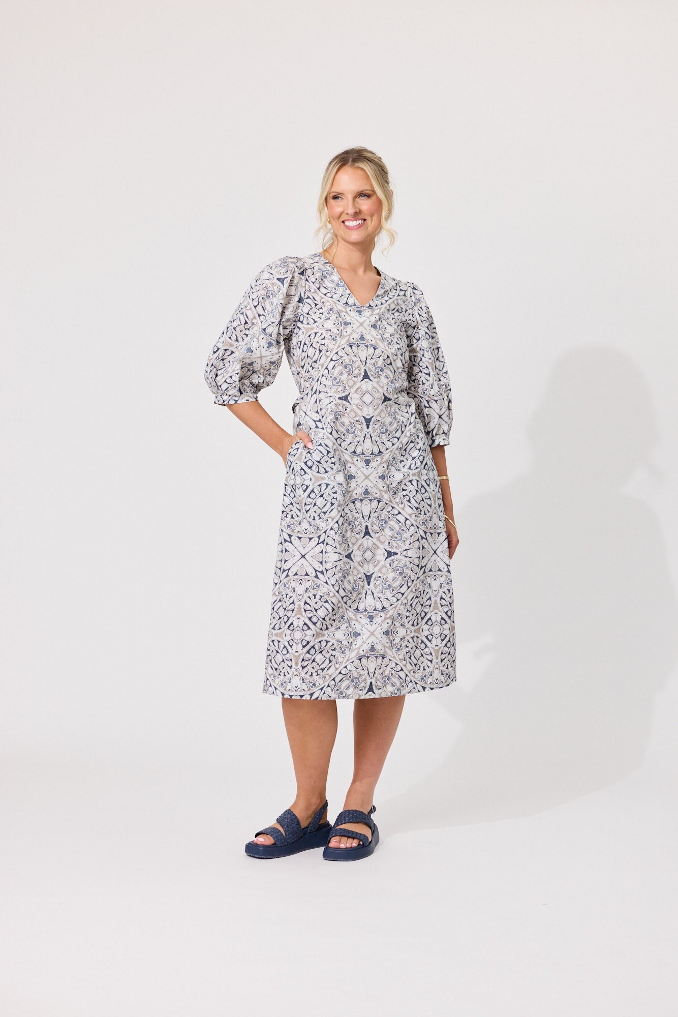 A woman modeling a knee-length dress with a blue and white floral pattern, featuring puff sleeves and a V-neckline, standing against a plain light gray studio background.