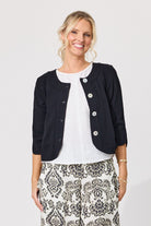 A smiling blonde woman models a short black cropped jacket with silver buttons over a white top and black-and-white patterned pants, shot against a light gray studio background.