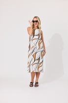 A woman modeling a sleeveless midi dress with a white background and an abstract brown and black leaf-like pattern, paired with black slide sandals and sunglasses, against a plain white studio background.