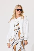 A woman modeling a white button-up shirt jacket over a white, black, and tan abstract print dress, wearing black sunglasses against a light gray studio background.