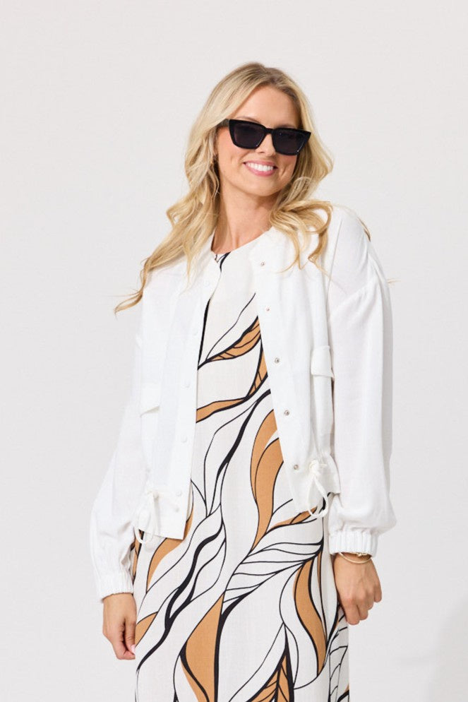 A woman modeling a white button-up shirt jacket over a white, black, and tan abstract print dress, wearing black sunglasses against a light gray studio background.