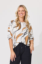 A smiling blonde woman wearing a loose-fitting white blouse with an abstract tan and black brushstroke pattern, paired with black pants, against a plain light gray studio background.