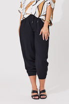 A model wearing black cropped jogger pants with a drawstring waist, paired with a patterned blouse and black sandals, shown from the waist down against a plain white studio background.